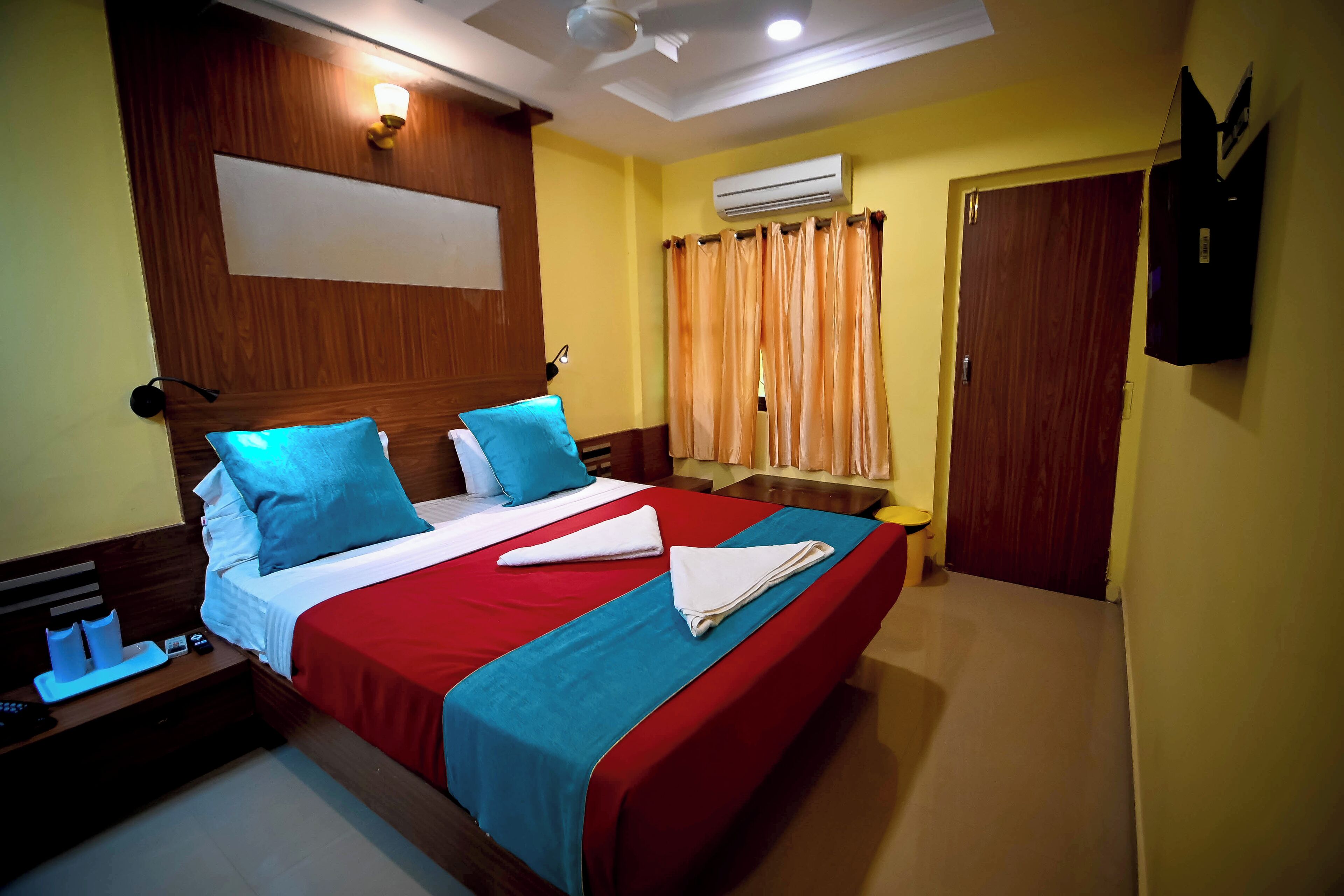 A hotel room with a large bed, two pillows, a red and blue bedspread, a nightstand with a remote control, and an air conditioning unit on the wall.