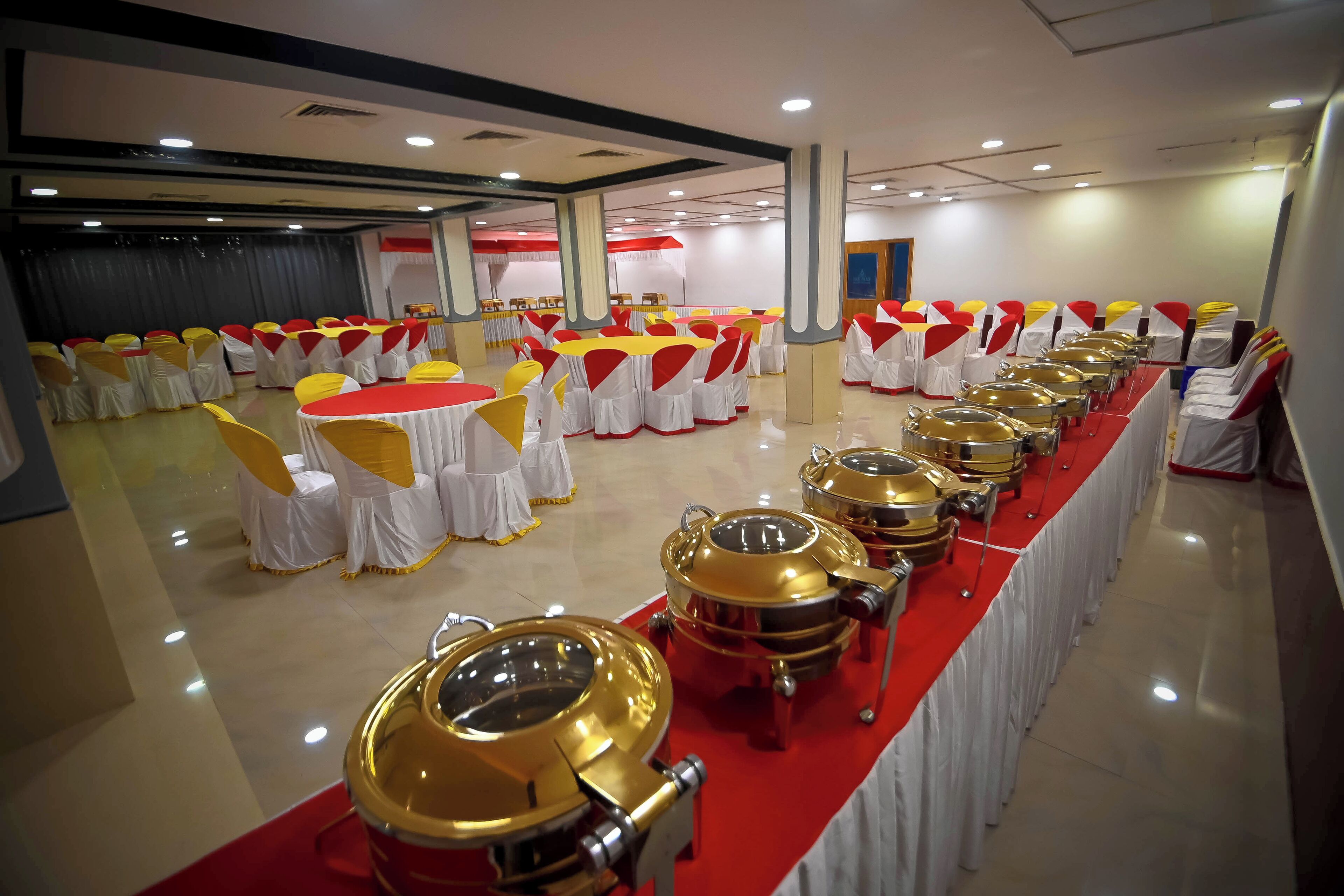 A banquet hall with rows of round tables covered with white tablecloths and yellow sashes, and gold buffet trolleys along the walls.