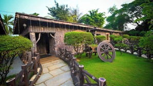 A rustic wooden building with a stone pathway leading to the entrance, surrounded by lush greenery and large wooden wheels. - Benzy Palace Resort (Bhatkal)