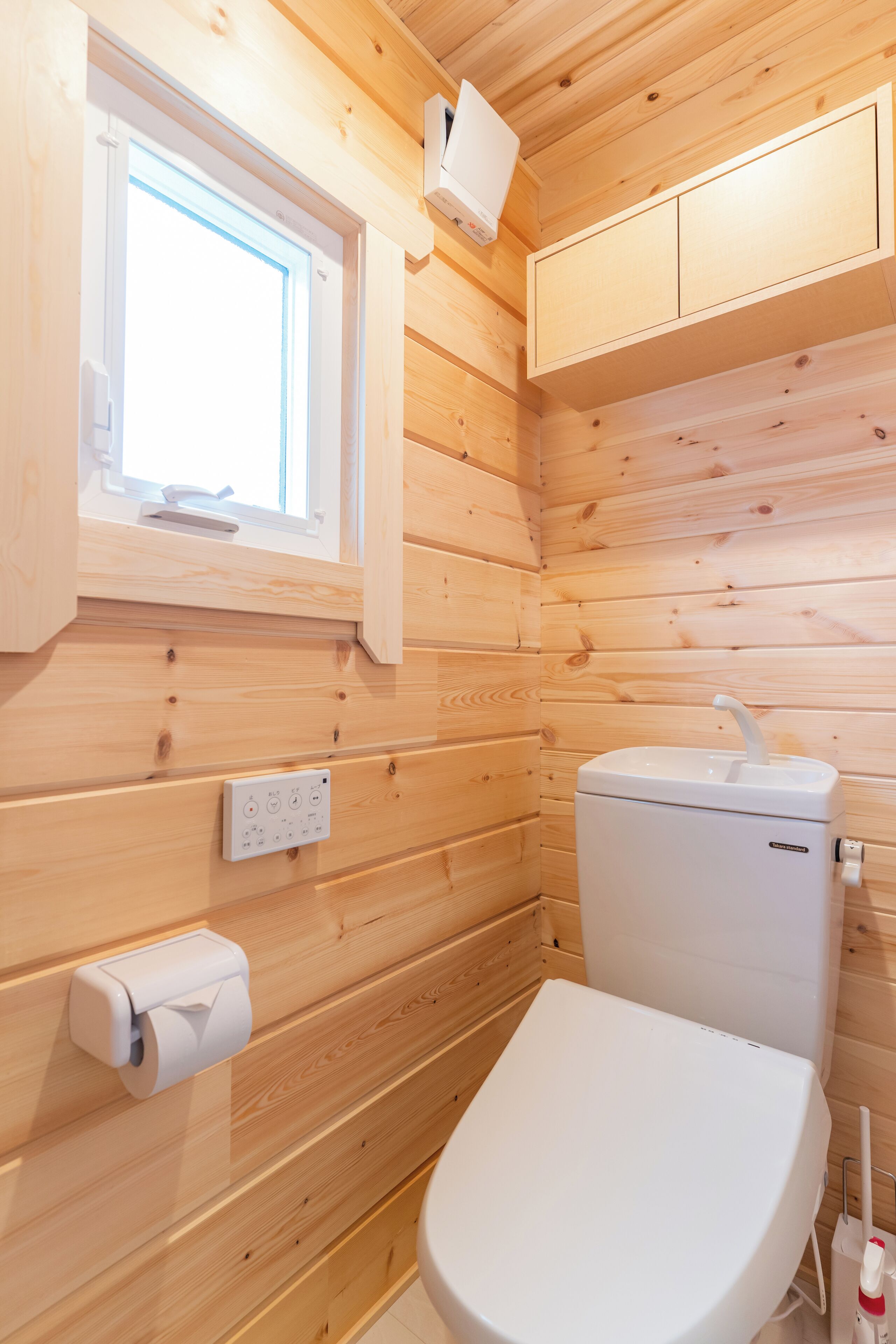 A wooden-paneled bathroom with a toilet, a window, and a wall-mounted electrical outlet.