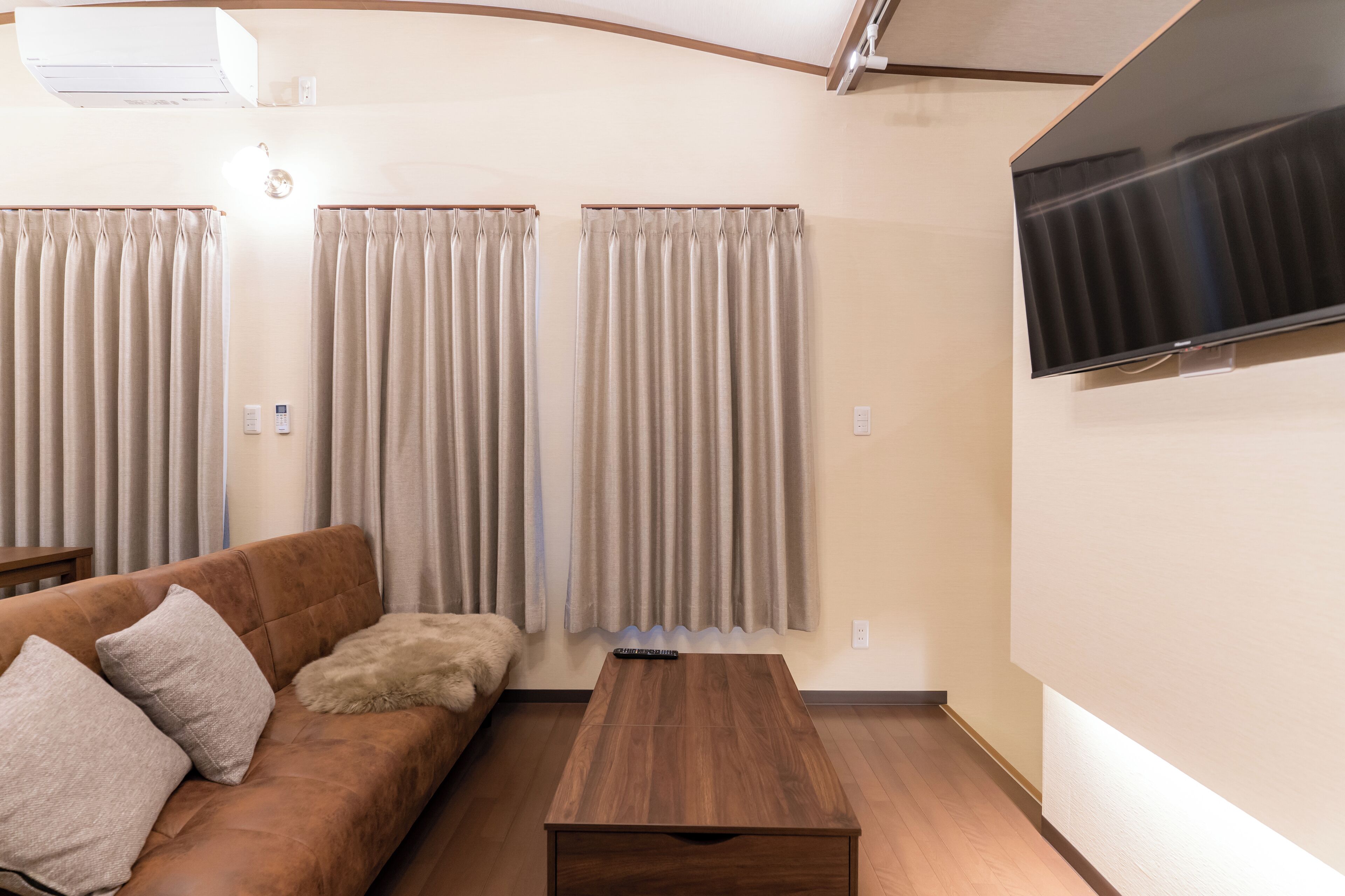 Ein Wohnzimmer mit braunem Sofa, beige farbenen Vorhängen, einem Holztisch und einem Wand-Fernseher.