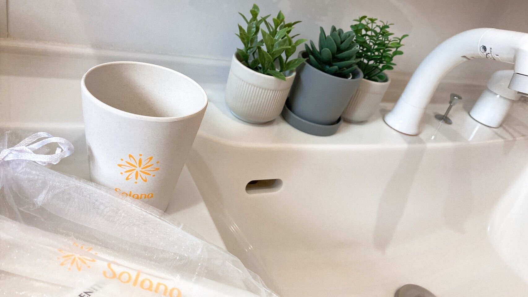 A bathroom sink with a white faucet, a white cup with orange text, and three potted plants.