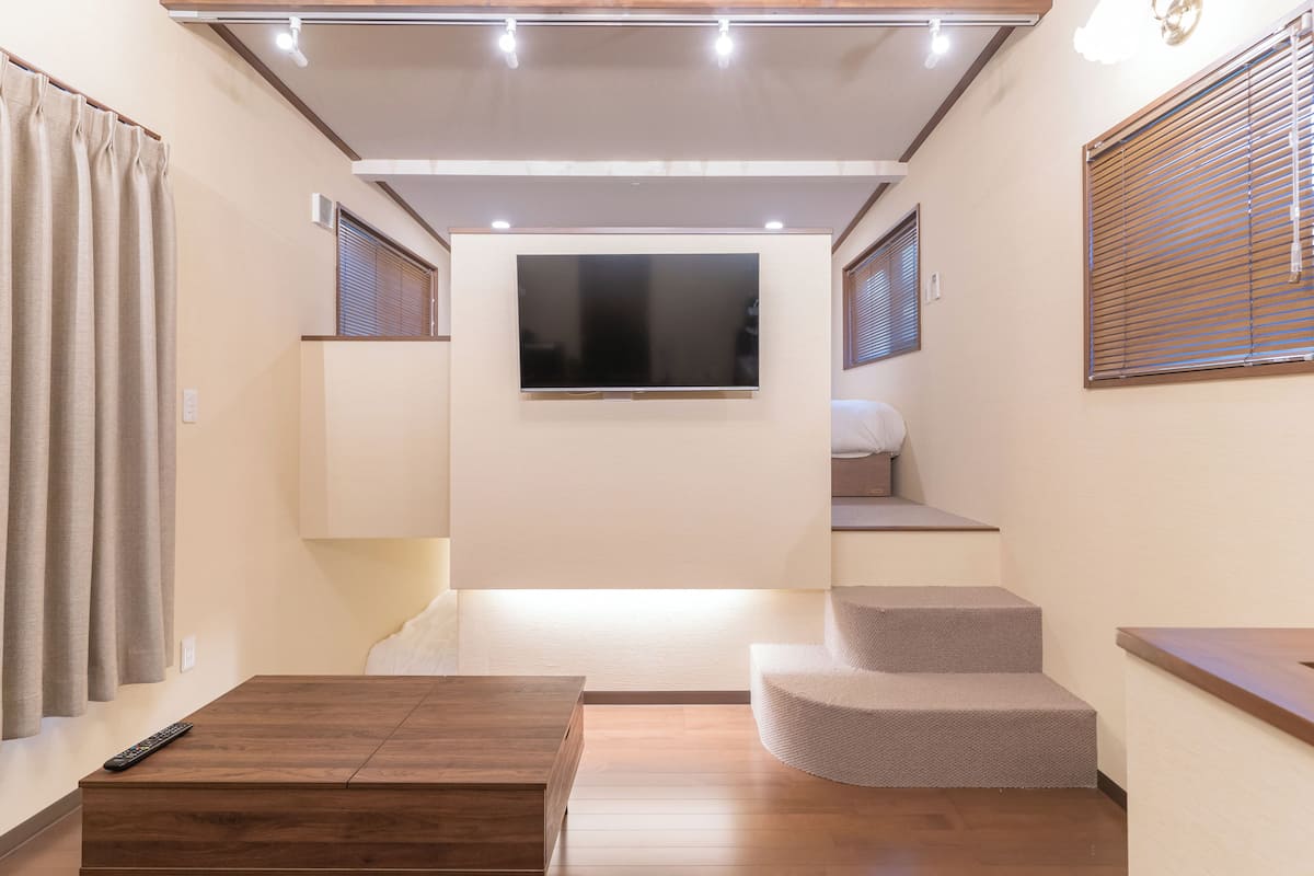 Una habitación de hotel compacta con televisor de pantalla plana montado en la pared, una mesita de madera, un banco empotrado y una escalera que lleva a un nivel superior.