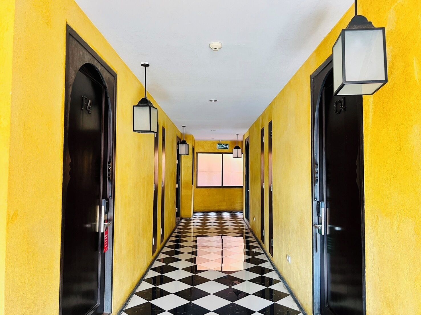 Un pasillo con paredes amarillas y piso a cuadros en blanco y negro que conduce a una habitación con ventana.