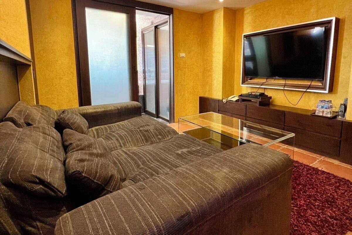 A modern living room with a large sectional sofa, a glass coffee table, a flat-screen TV mounted on the wall, and a sliding glass door leading to a balcony.