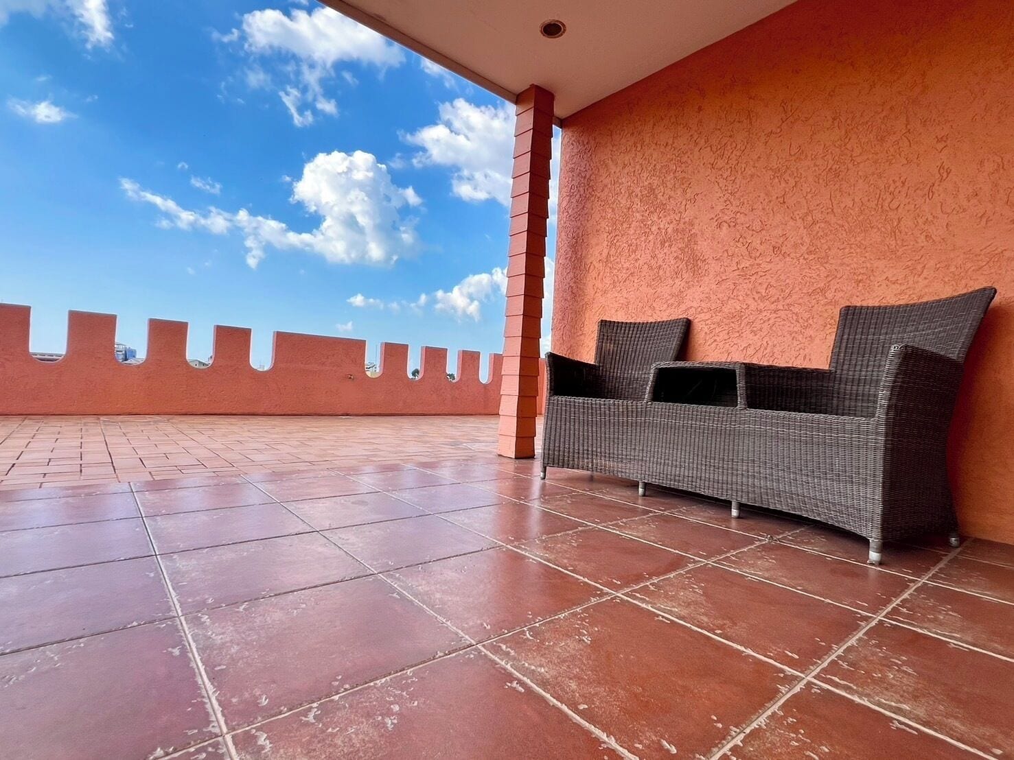 A covered patio area with a wicker sofa and a tiled floor.
