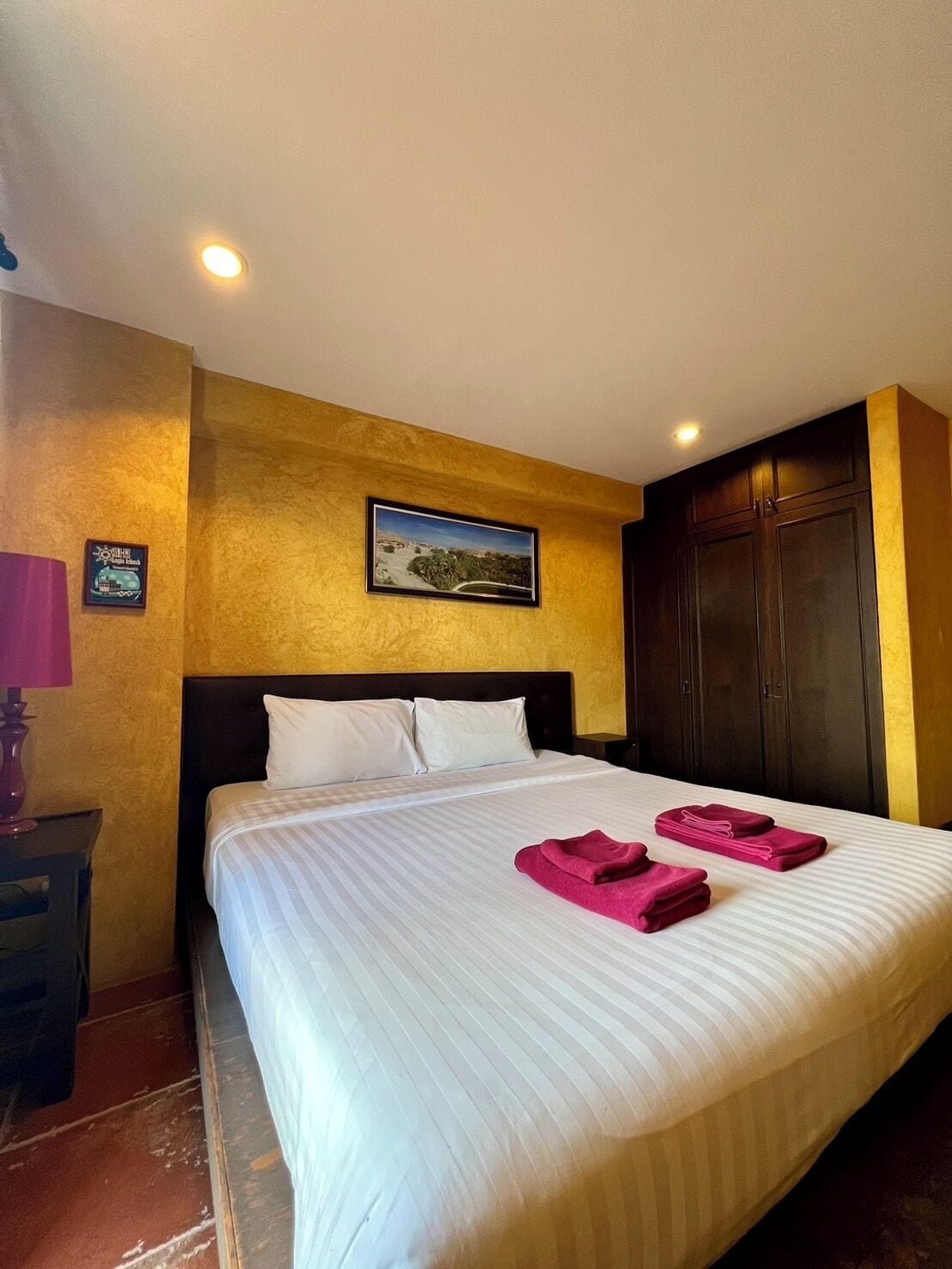 Una habitación de hotel con una cama grande, una mesita de noche, una lámpara, un cuadro en la pared y un armario.