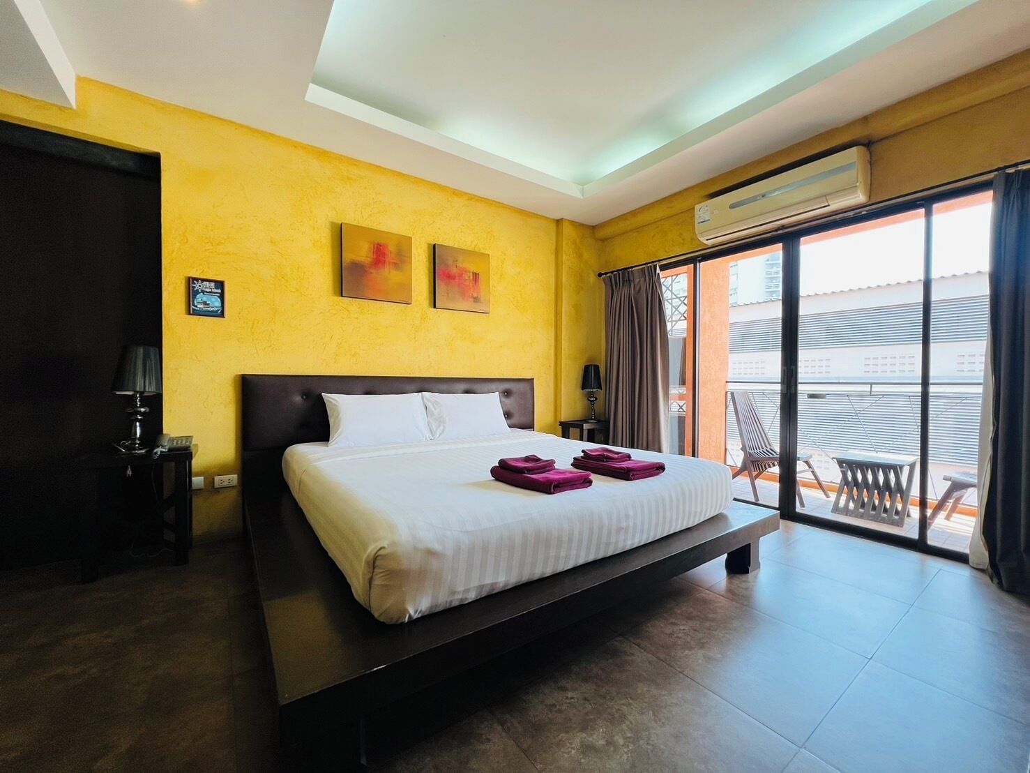 A hotel room with a large bed, a nightstand, an air conditioning unit, and a view of the outside balcony.