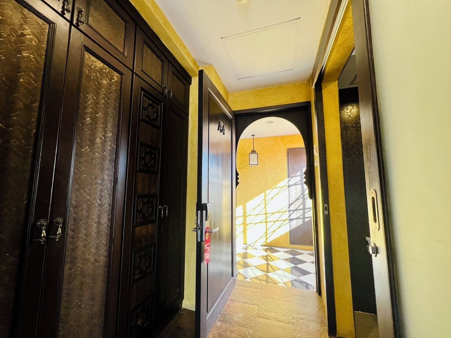 A hallway with dark wooden doors, a yellow wall, and a checkered floor leading to a staircase.