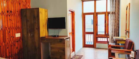 Quarto de hotel com teto de madeira, televisão sobre suporte de madeira, cama, escrivaninha e área de estar com cadeiras.