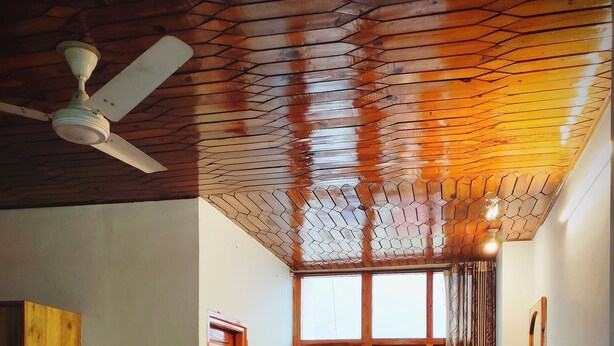 A hotel room with a wooden ceiling, a bed, a desk with a chair, a television, and a large window with curtains. - HimalayanHamlet ValleyView (Kullu)