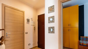 Un pasillo con una puerta de madera, un teléfono y un reloj en la pared. Se ve un armario amarillo y un sofá colorido en la habitación contigua.