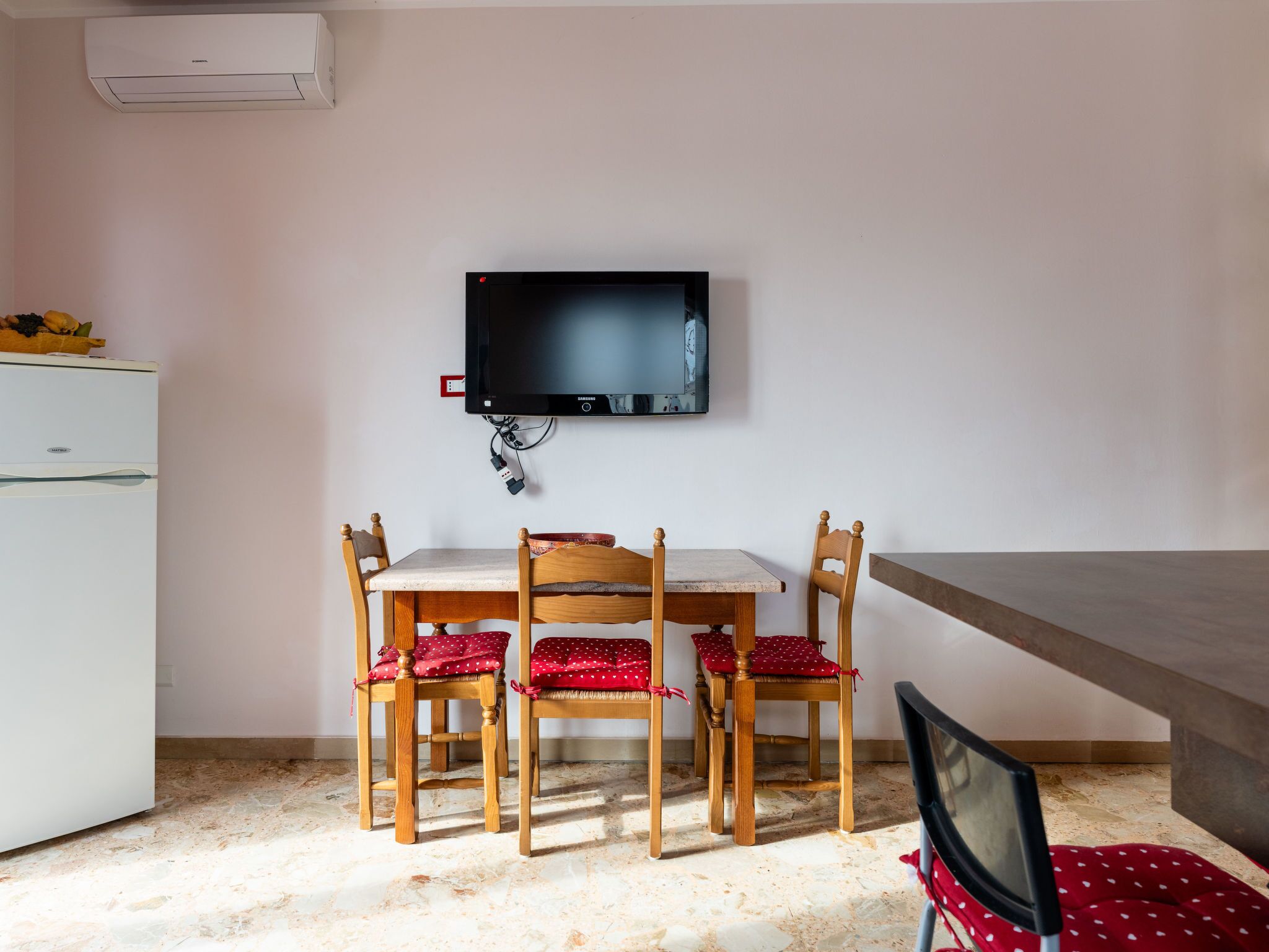 A small dining area with a white table, four wooden chairs with red cushions, a flat-screen TV mounted on the wall, and a white refrigerator.