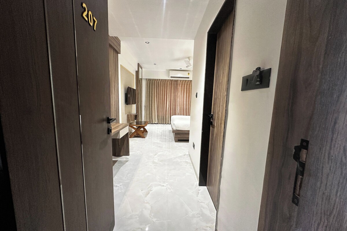 A hotel hallway with a marble floor, a wooden door labeled '207', a bed with a white headboard, and a television mounted on the wall.