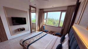 A modern hotel room with a large bed, a flat-screen TV mounted on the wall, a small seating area with a table and chairs, and a balcony with a view of the city and mountains. - Hotel Kushala Grand, Dombivali (Ambernath)