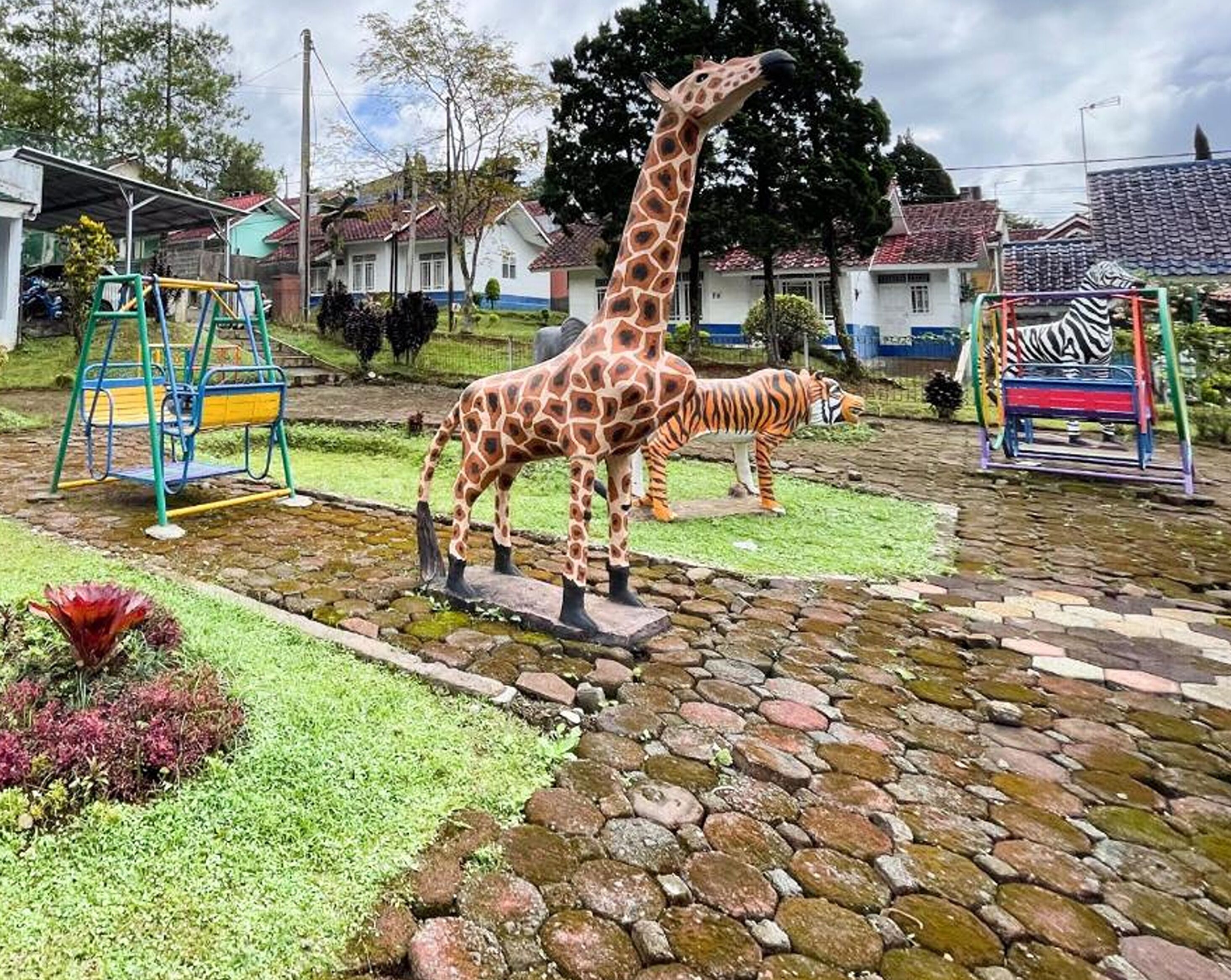 A playground with a giraffe and zebra sculpture, surrounded by houses and greenery.