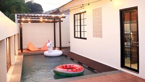 A pool area with a white swan float and a watermelon float. There is a pergola with string lights and a wooden deck.