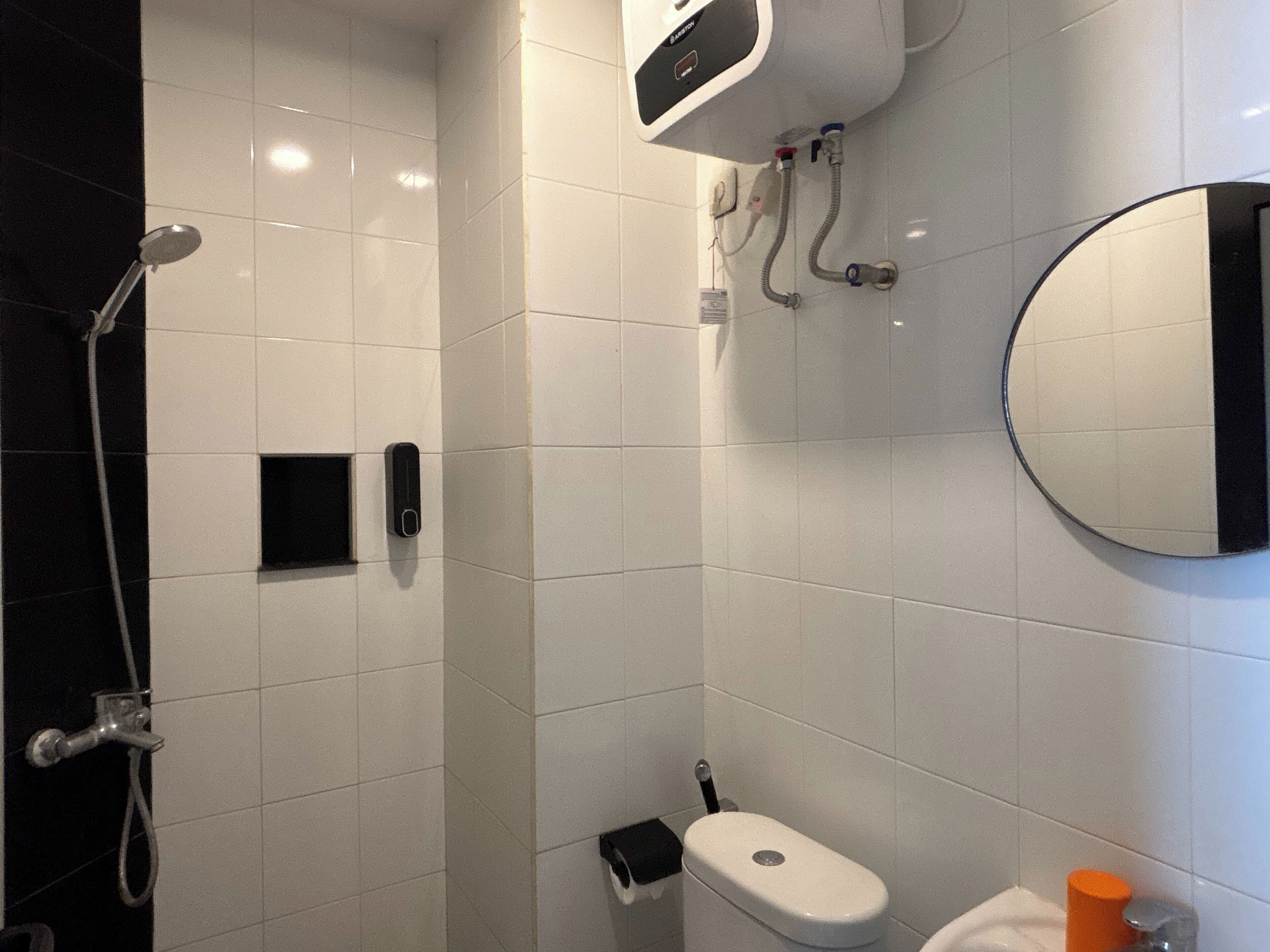 Une salle de bain moderne avec des carreaux blancs, des toilettes, une douche équipée d’un pommeau de douche amovible et un miroir fixé au mur.
