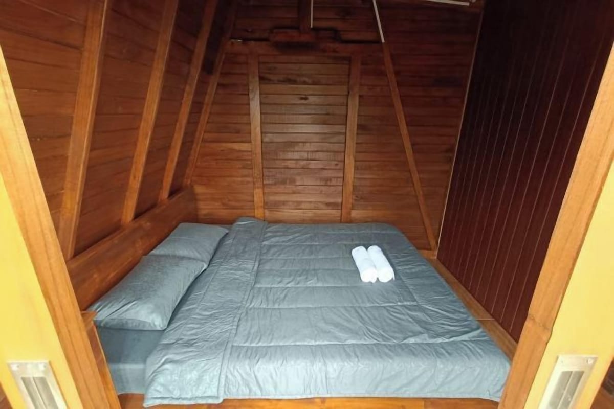 A small, narrow room with a wooden ceiling and walls, a single bed with a grey mattress and pillows, and a pair of rolled-up towels on the bed.