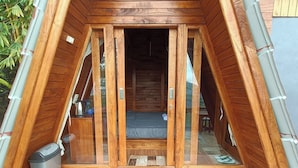 Een houten A-frame constructie met een bed, een kleine tafel, een prullenbak en een raam.