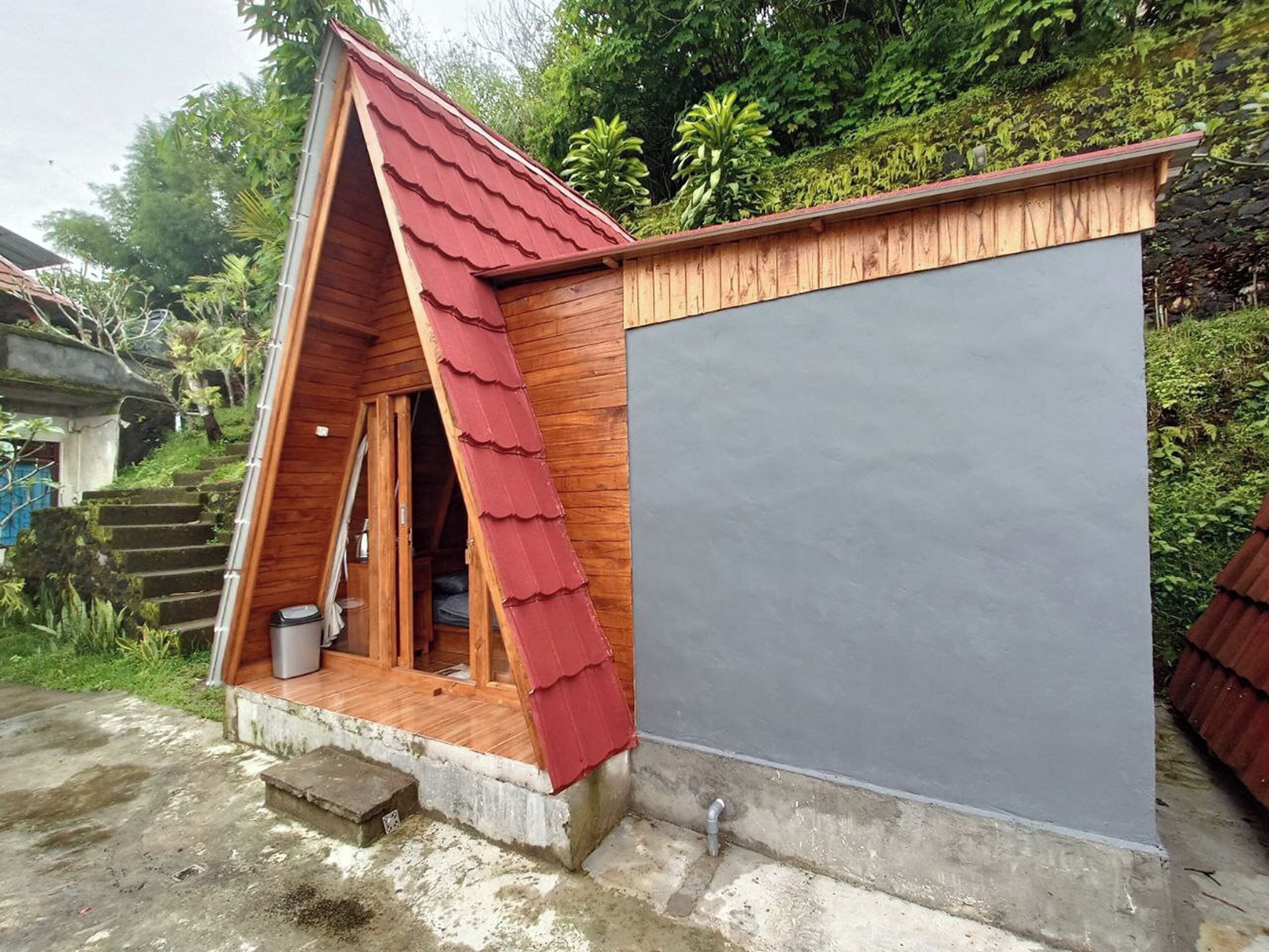 A small wooden A-frame structure with a red roof, a grey wall, and a concrete floor.