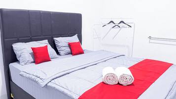 Uma cama bem arrumada com cabeceira cinza, almofadas cinzas e vermelhas, e um cobertor cinza e vermelho. Existem duas toalhas enroladas sobre a cama.