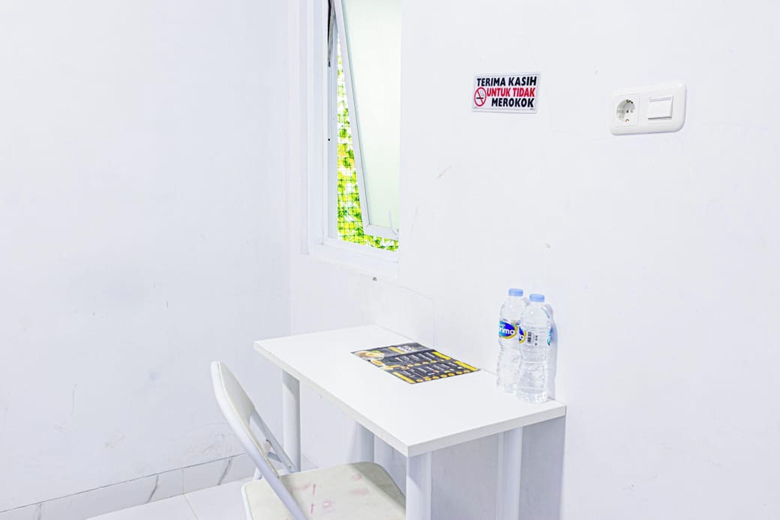 Een kleine witte kamer met een raam, een tafel met een tijdschrift en waterflesjes, en een bordje aan de muur.