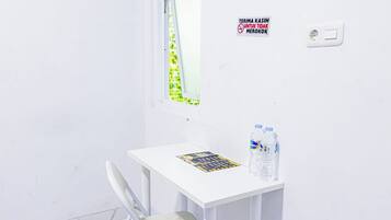 Een kleine witte kamer met een raam, een tafel met een tijdschrift en waterflesjes, en een bordje aan de muur.
