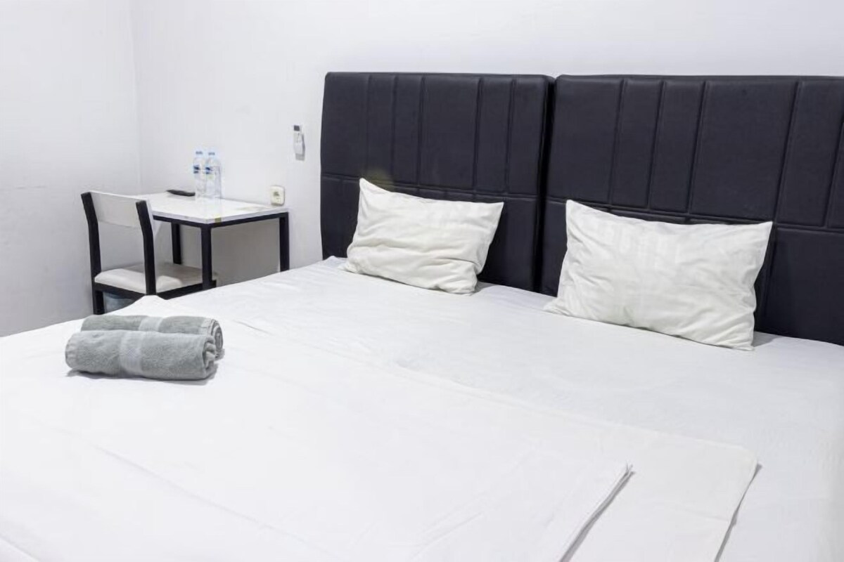 A neatly made bed with white linens and pillows, a small table with a chair, and a water bottle on the table.