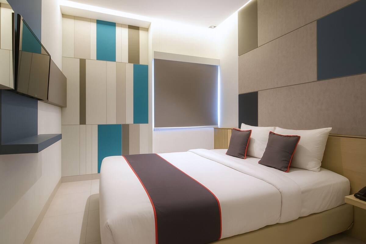 A modern hotel room with a large bed, a flat-screen TV mounted on the wall, and a window with blinds.
