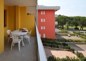 A balcony with a white table and chairs, a yellow wall, and a view of a landscaped garden with trees and a building in the background. - Studio in excellent location (Bibione)