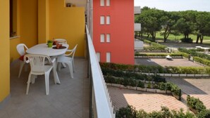 Un balcone con tavolo e sedie bianche, una parete gialla e vista su un giardino curato con alberi e un edificio sullo sfondo.