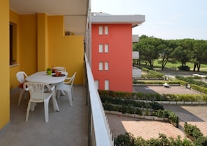A balcony with a white table and chairs, a yellow wall, and a view of a landscaped garden with trees and a building in the background. - Well-located flat with balcony (Bibione)