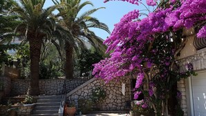 Een stenen trap leidt naar een garage, omgeven door weelderige begroeiing en paarse bougainvillea bloemen.