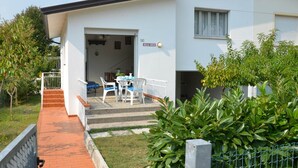 Une maison moderne à deux étages, avec une façade blanche, un patio couvert agrémenté de mobilier d’extérieur et un escalier apparent menant à l’entrée.