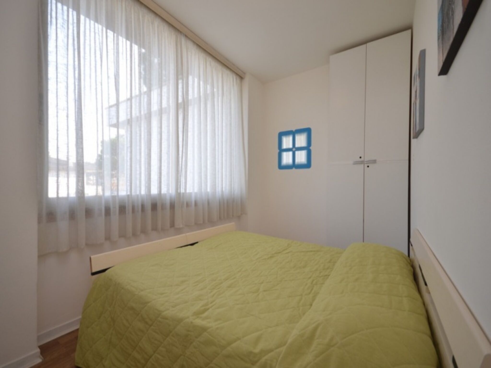 A room with a bed, a window with sheer curtains, a door with a blue window decal, and a wall-mounted picture frame.
