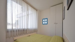 Una habitació amb un llit, una finestra amb cortines primes, una porta amb un adhesiu de finestra blau i un quadre penjat a la paret.