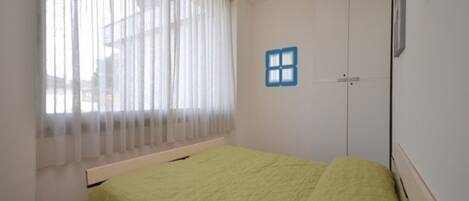 A room with a bed, a window with sheer curtains, a door with a blue window decal, and a wall-mounted picture frame.
