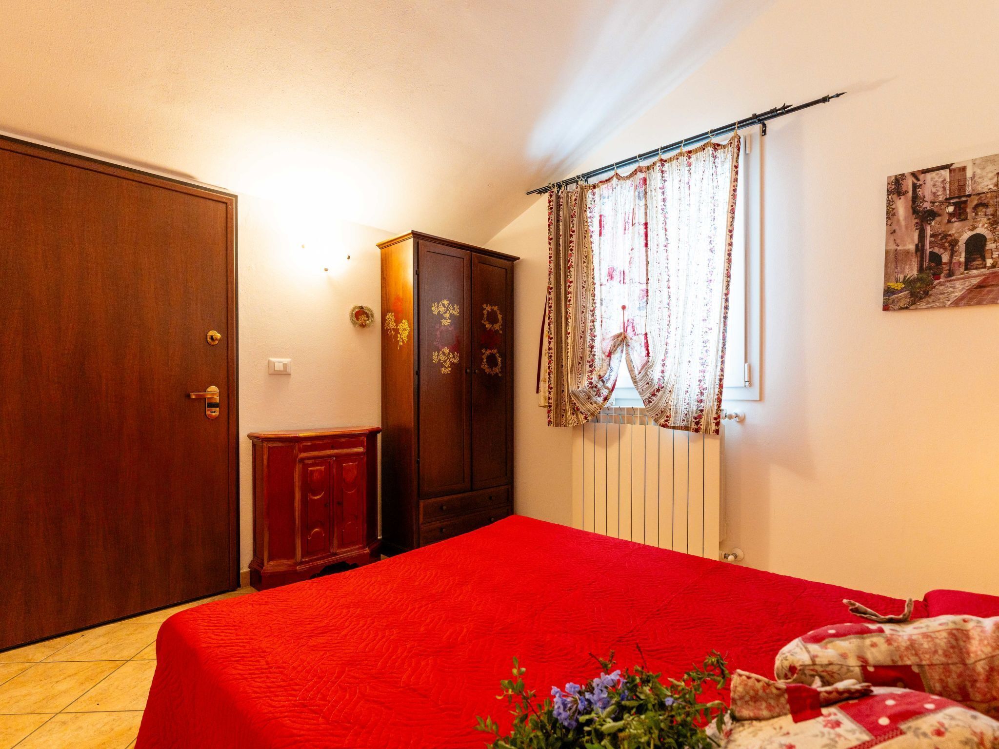 A bedroom with a red bedspread, wooden wardrobe, and a window with floral curtains.