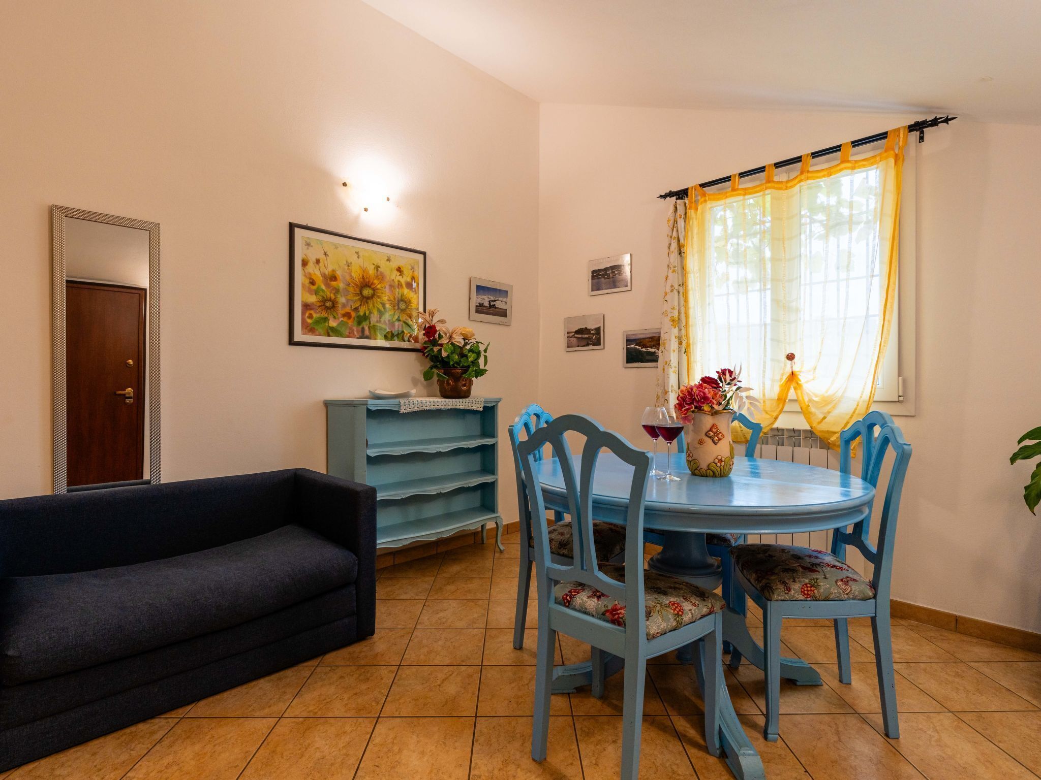 A dining area with a blue table and chairs, a dark sofa, a blue cabinet, and a window with yellow curtains.