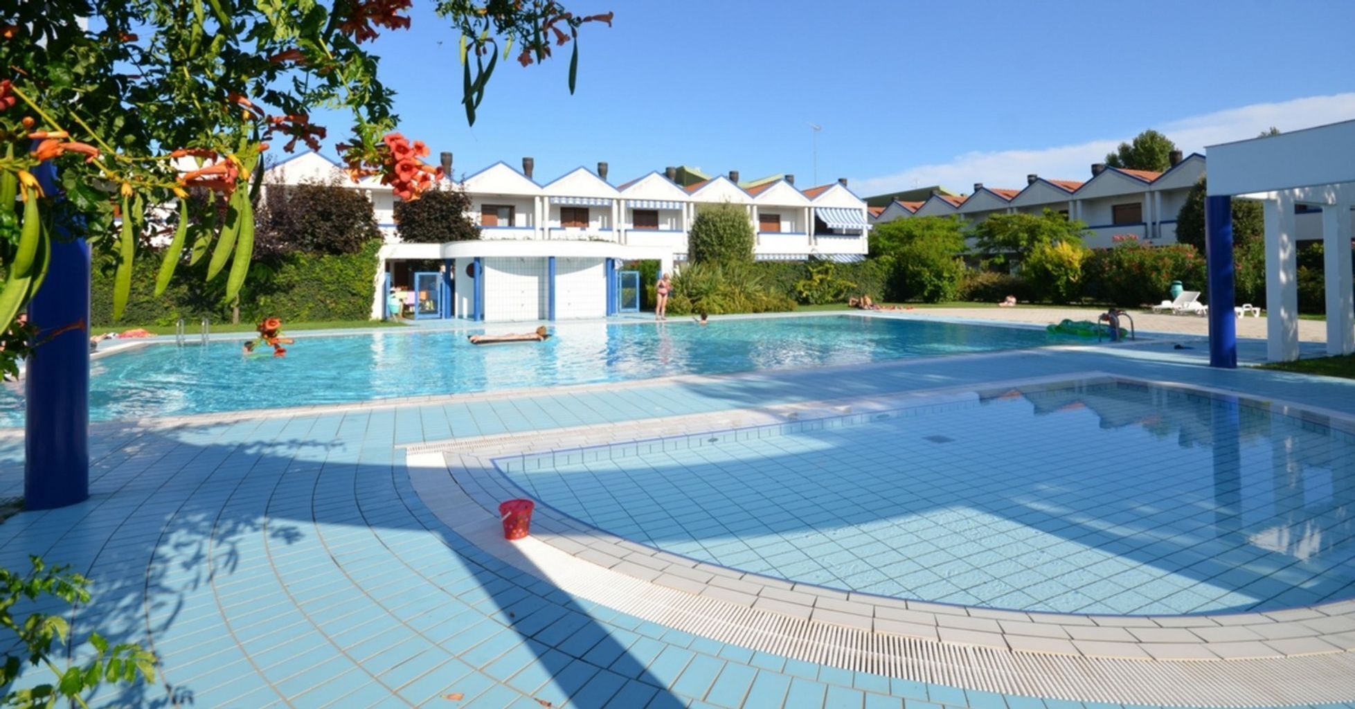 Piscina com piso de azulejos, cercada por um edifício com várias varandas e céu azul límpido.