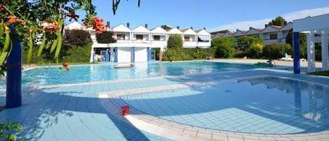 Una piscina con suelo de baldosas, rodeada por un edificio con varios balcones y un cielo azul despejado.