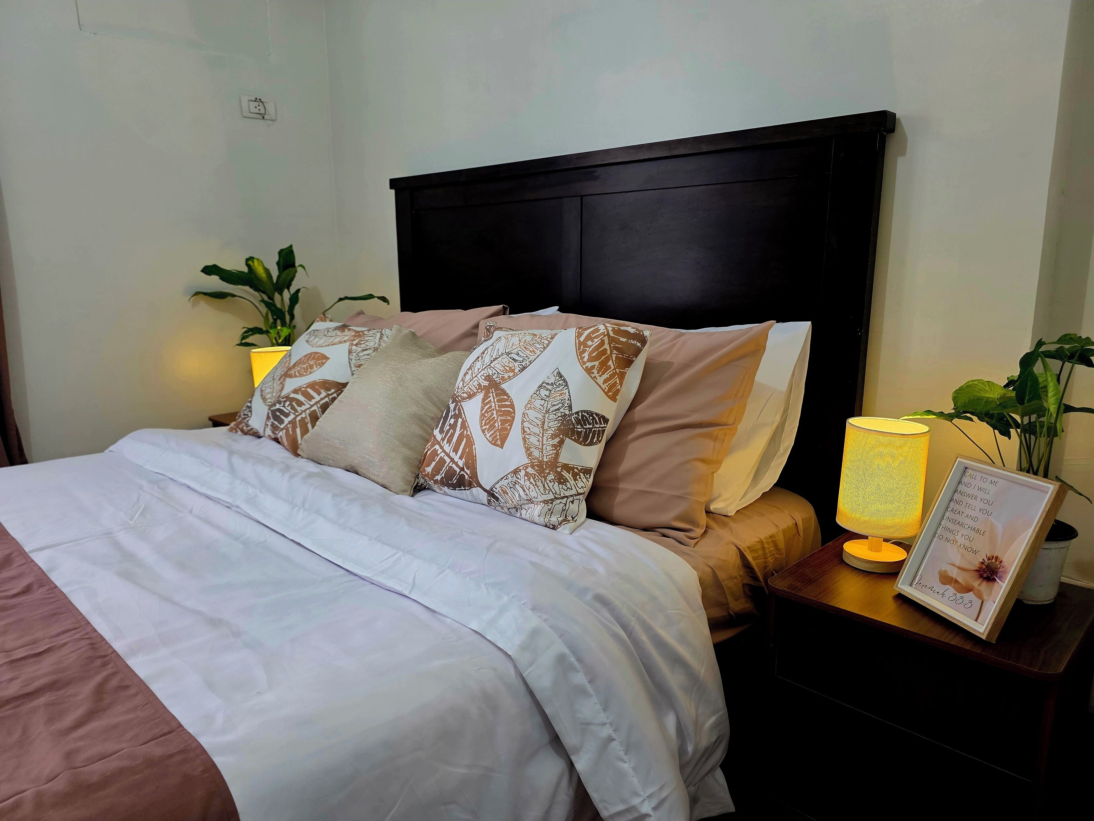 A neatly made bed with patterned pillows, a bedside table with a lamp, and a framed quote on the wall.