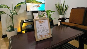 Un área de recepción con un televisor mostrando una escena de playa, un cartel de bienvenida y varias plantas en macetas.