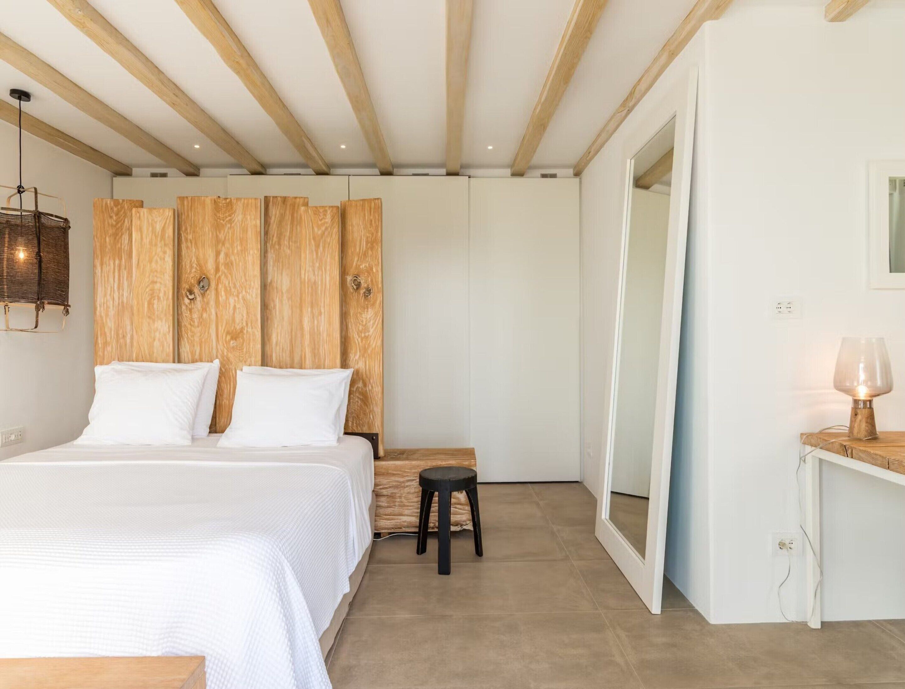 A bedroom with a bed, wooden headboard, a small stool, a bedside table with a lamp, and a mirror.