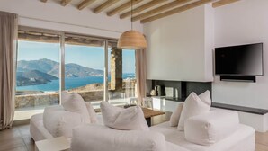Un salon moderne avec un canapé blanc, un téléviseur fixé au mur et une grande fenêtre offrant une vue panoramique sur la mer et les montagnes.