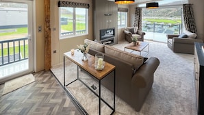 A spacious living area with a sofa, coffee table, and TV, large windows with a view of the outdoors, and a wooden ceiling beam.