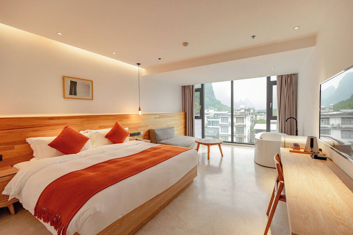 A modern hotel room with a large bed, a desk, and a view of a cityscape through large windows.