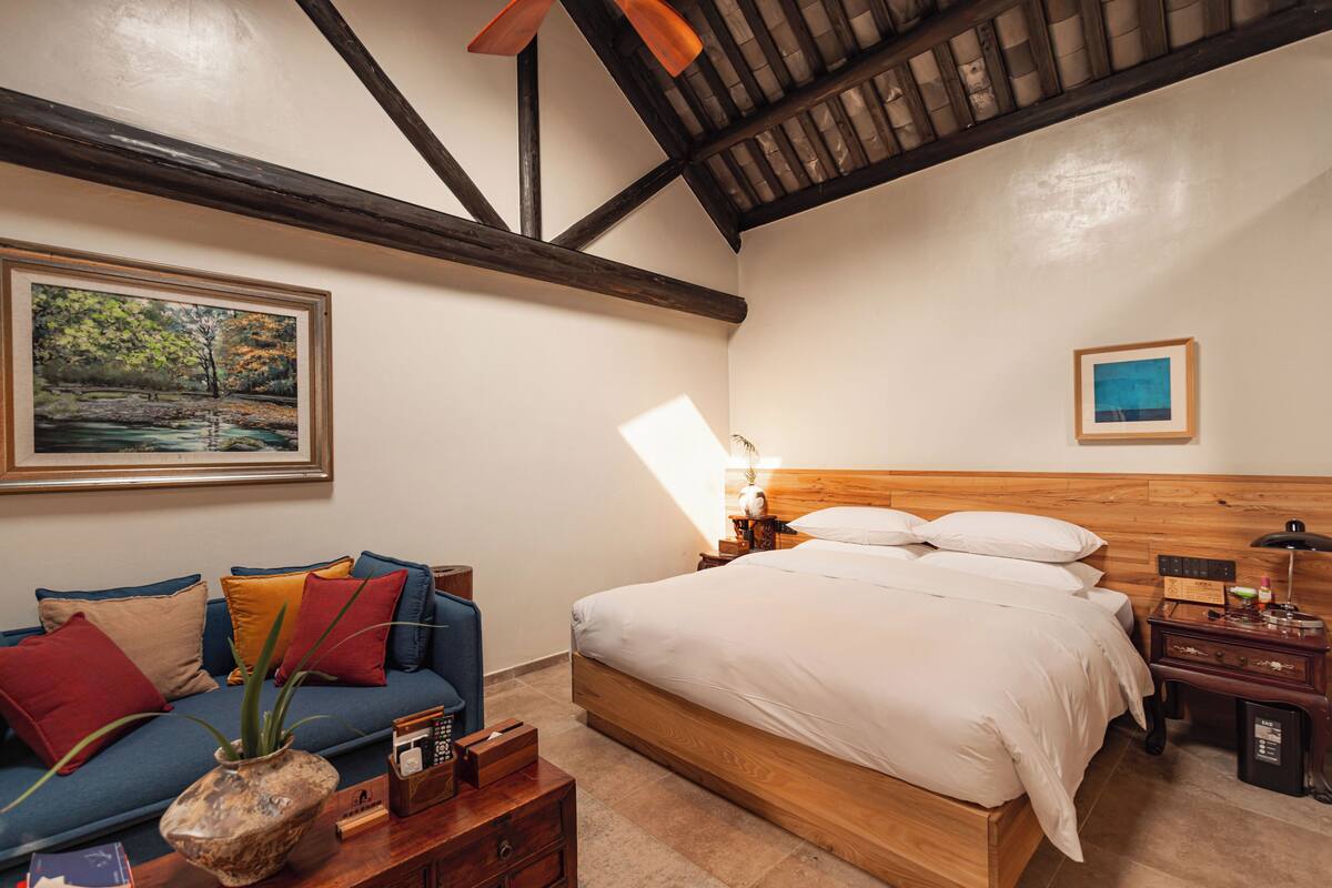 A bedroom with a wooden bed, a blue sofa with colorful cushions, a nightstand with a lamp, and a framed painting on the wall.