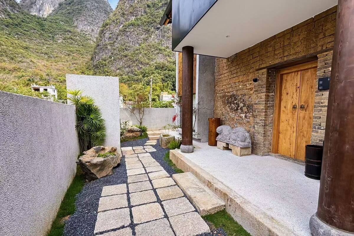 A modern building with a stone pathway leading to a wooden door, set against a mountainous backdrop.